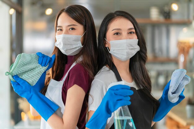 Photo portrait of asian waitress with apron staff wearing protection rubber glove face mask protection hand hold cleaning towel and alcohol spray disinfectant for protect infection coronavirus health ideas