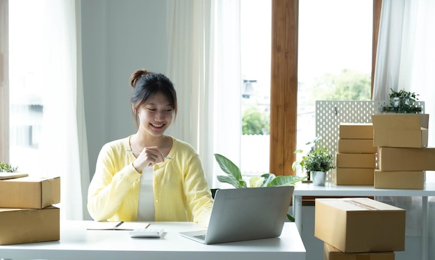 A portrait of Asian woman ecommerce employee sitting in the office full of packages on the table using a laptop and calculator for SME business ecommerce technology and delivery business