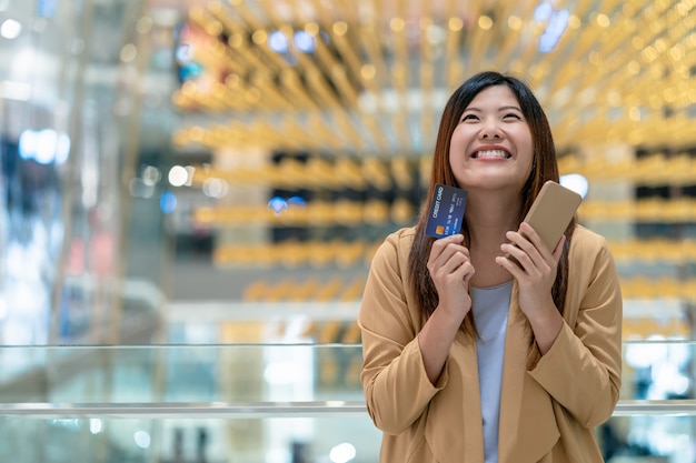 Felicità asiatica della donna del ritratto quando tengono la carta di credito e il telefono cellulare per acquisto online nel grande magazzino