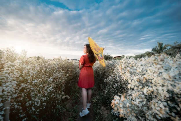 Photo portrait of asian woman happy traveler with red dress hold yellow umbrella enjoying in white blooming or white margarita flowe field in the garden of in chiang mai,thailand,travel relax vacation