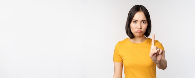 Photo portrait of asian woman looking serious and angry showing stop prohibit gesture taboo sign forbidding smth standing in yellow tshirt over white background