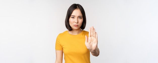 Portrait of asian woman looking serious and angry showing stop prohibit gesture taboo sign forbidding smth standing in yellow tshirt over white background