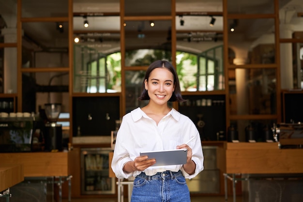 カフェの入り口の前にタブレットを持って立っているアジアの女性マネージャーの肖像画がゲストを歓迎します