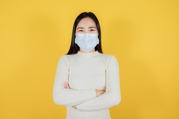 Portrait of Asian woman in medical face mask to protect Covid-19 (Coronavirus) wears cream sweater standing crossed arms on yellow background