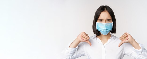 Portrait of asian woman in medical face mask showing thumbs down with disappointed tired face expres