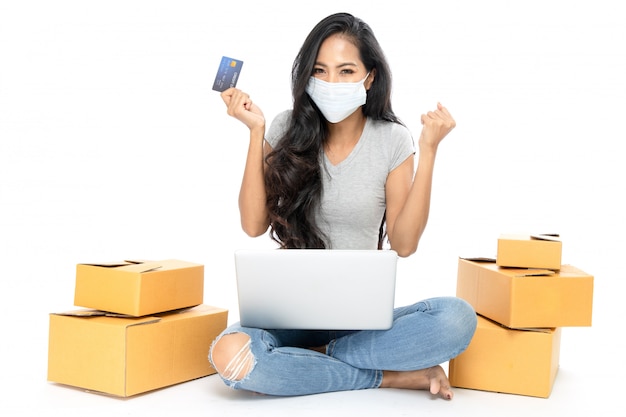 Portrait of an Asian woman sits on the floor with a lot of boxes on the side. She is holding a credit card to shop online. She wears medical flu mask. Isolated on white background