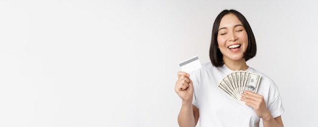 Portrait of asian woman smiling holding credit card and money cash dollars standing in tshirt over w...