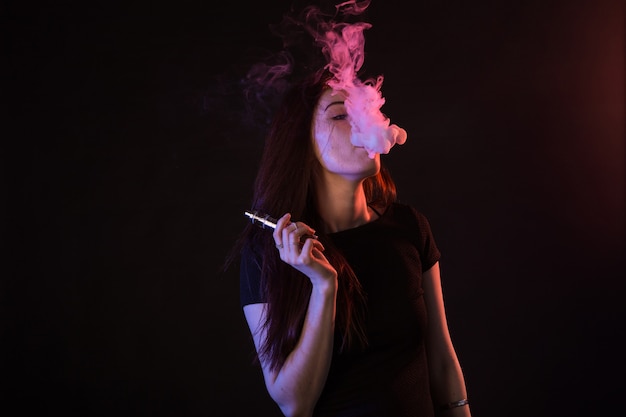 검정색 배경에서 네온 불빛에 아시아 여자 흡연 vape 또는 ecigarette의 초상화.