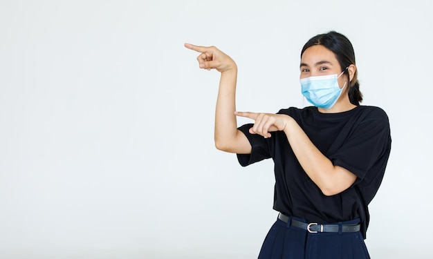 Portrait Asian woman wearing a face mask poses, pointing advertisement on white background, blank copy space with isolated white background cutout.