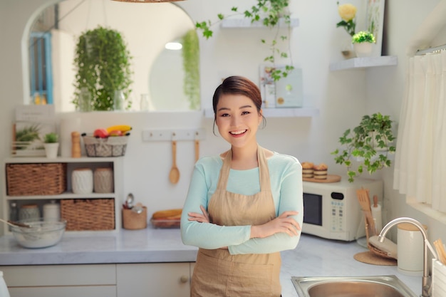 Портрет азиатской молодой счастливой красивой женщины, улыбающейся стоя, скрещивая руки на кухне