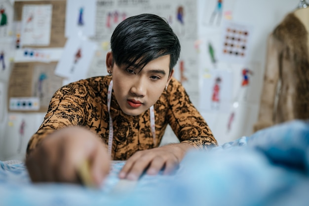 Портрет азиатского молодого мужчины портного вырезает кусок ткани за столом в студии моды.