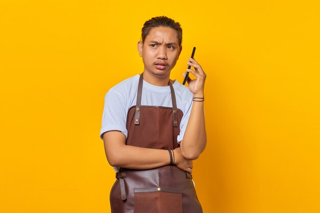 Portrait of asian young man wearing apron looking angry while talking on smartphone