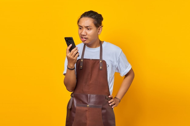 Portrait of Asian young man wearing apron looking angry while talking on smartphone