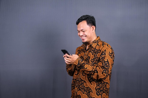 Portrait of asian young man wearing batik using cellphone on gray background