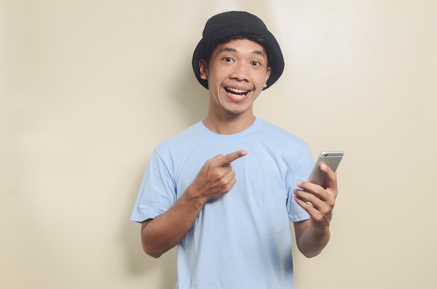 Портрет азиатского молодого человека в синей футболке и черной шляпе, указывающего на телефон на изолированном фоне