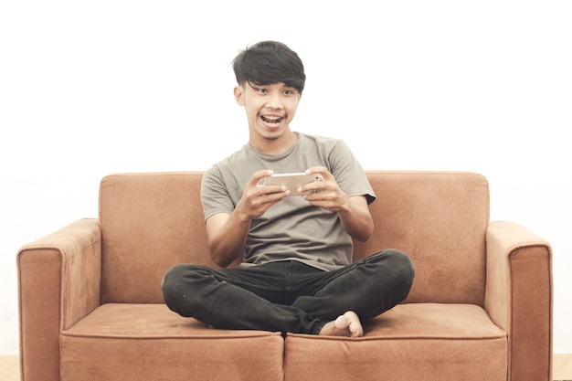 彼の携帯電話でゲームをしているソファに座っている灰色のTシャツを着ているアジアの若い男の肖像画