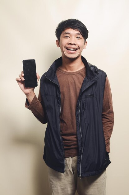 Портрет азиатского молодого человека с взволнованным выражением лица, показывающего экран телефона в коричневой футболке и черном жилете на заднем плане