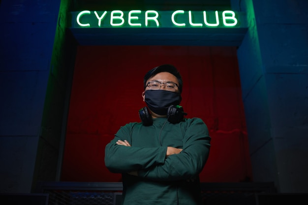 Foto ritratto di giovane programmatore asiatico in maschera in piedi con le braccia incrociate vicino al club di computer e che guarda l'obbiettivo