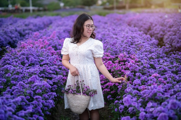 白いドレスを着たアジアの若い女性の幸せな旅行者のポートレートは、チェンマイの自然園で白い花または紫のミカエルマス・デイジーの花畑を楽しみ、タイ旅行でリラックスした休暇を過ごします