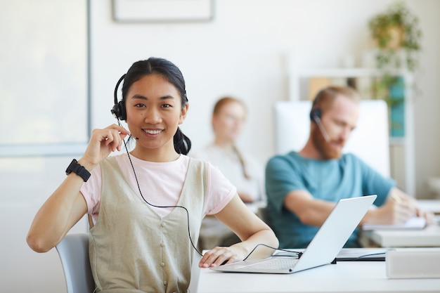 Portrait of Asian young woman in headset smiling while working at her workplace with laptop