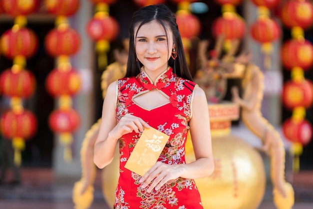 Портрет азиатской молодой женщины в красном традиционном китайском чонсаме, держащей желтые конверты с китайским текстом «Благословения», написанными на нем - удача на фестивале китайского Нового года