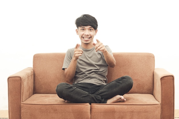 Портрет азиатской молодежи в серой футболке, сидящей на диване, указывая вперед