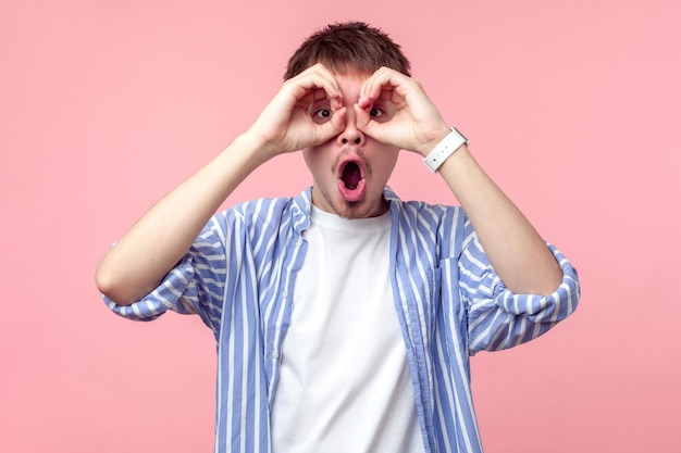 Portrait of astonished curious brown-haired man with small beard and mustache in casual shirt looking through fingers gesturing binoculars, shocked gaze. indoor studio shot isolated on pink background