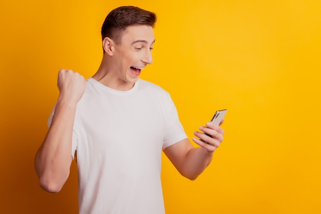 Portrait of astonished excited guy hold smartphone raise fist winner concept on yellow background