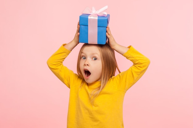 Photo portrait of astonished little girl holding gift box on head with expression of much surprise and amazement, shocked by birthday present, holiday bonus. indoor studio shot isolated on pink background