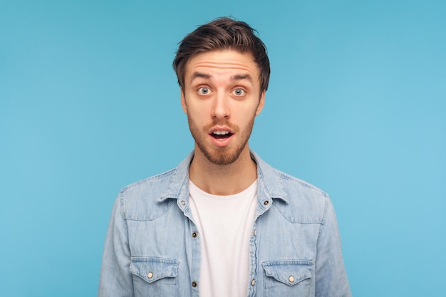 Portrait of astonished man wearing denim shirt standing with mouth open in surprise has shocked expression hears unbelievable news Indoor studio shot isolated on blue background