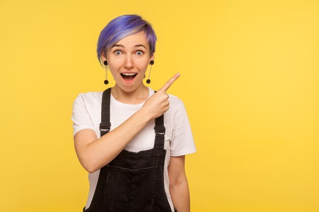 Portrait of astonished surprised fashionable hipster girl with violet short hair in denim overalls pointing to the side freespace for advertising copy space isolated on yellow background studio shot