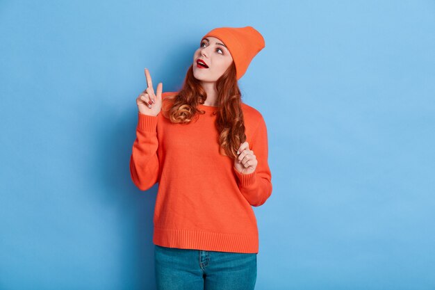 Portrait of astonished woman point index finger at copy space, being impressed, wears sweater, jeans and cap