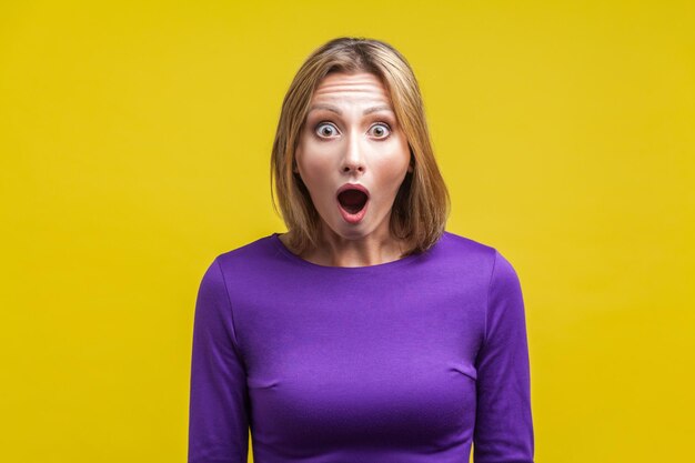 Portrait of astonished woman with widely open mouth in amazement and big eyes looking at camera, stunned shocked face. indoor studio shot isolated on yellow background