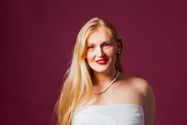 Portrait of astonishing blonde with loose hair on one side bare shoulers Beautiful pearl necklace on bare neck Woman with nude makeup and glossy red lips looking at the camera