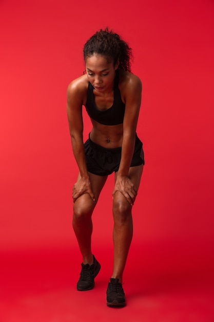 Portrait of athletic african american woman in black sportswear standing, isolated over red wall