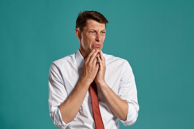 Portrait of an athletic brunet fellow with brown eyes, wearing in a classic white shirt and red tie. He is acting like he is afraid of something while posing in a studio against a blue background. Con