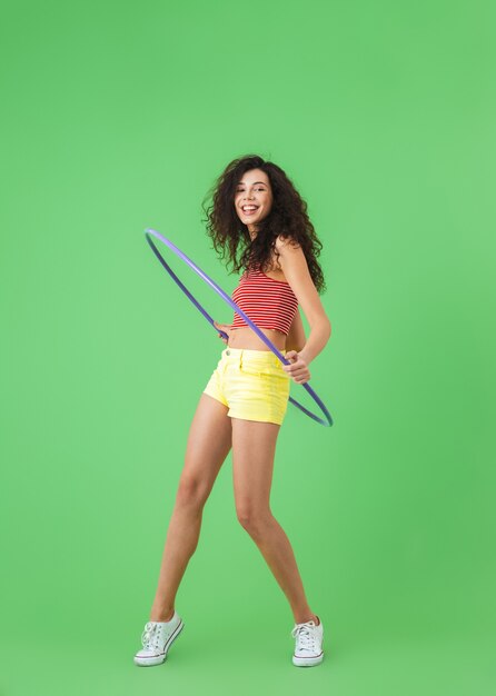 portrait of athletic woman 20s wearing summer clothes doing exercises with hula hoop during workout on green wall