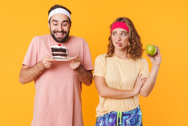 Portrait of athletic young couple wearing headbands choosing beetwen cake and apple isolated over yellow wall