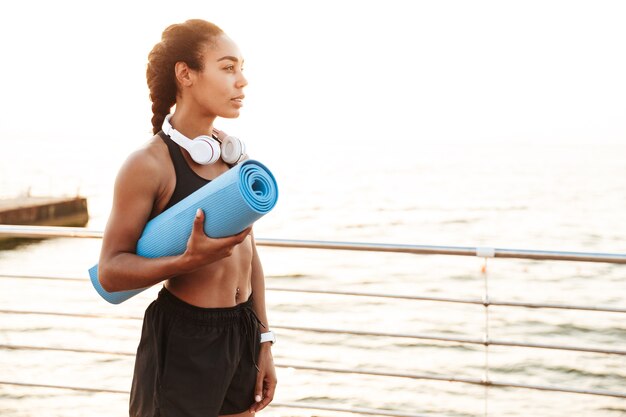 Portrait of athletic young woman in sportive clothes with headphones looking aside while standing with fitness mat by seaside in morning