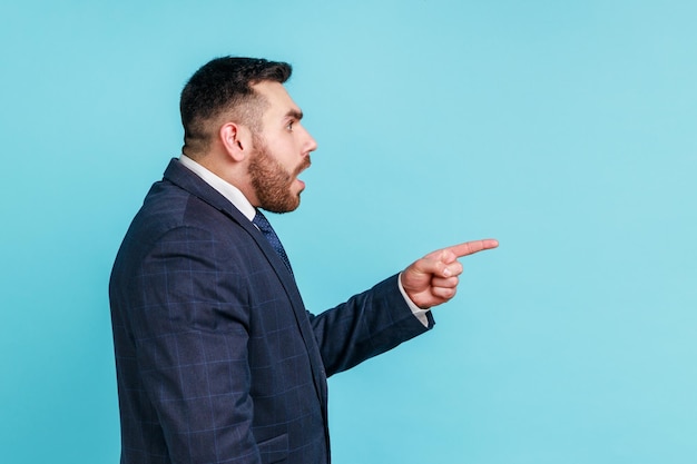 Photo portrait of attentive handsome bearded young man wearing official style suit having shocked facial expression pointing finger aside indoor studio shot isolated on blue background