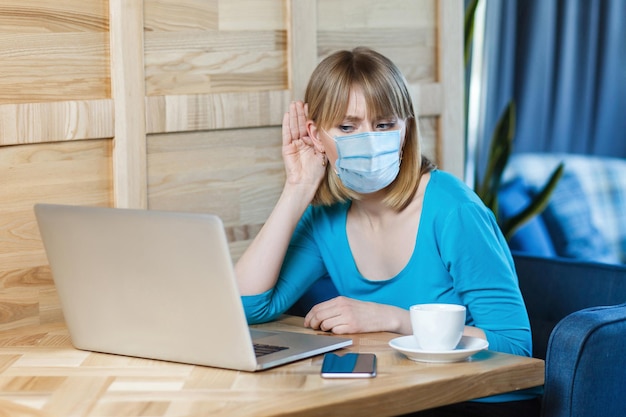 Il ritratto di una giovane donna attenta con maschera medica chirurgica è seduto e lavora su un laptop e sta cercando di ascoltare con attenzione al laptop. concetto di lavoro al chiuso, medicina e assistenza sanitaria.