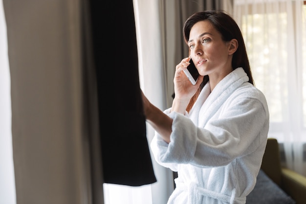 Portrait of attractive adult businesswoman in white bathrobe talking on cellphone while looking at her formal suit in apartment