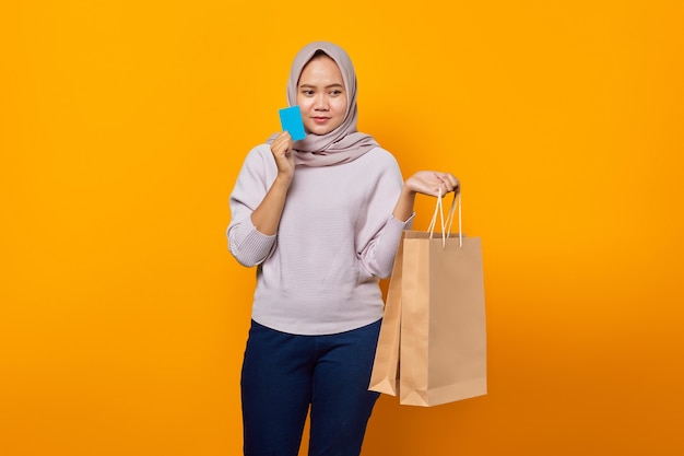 Portrait of attractive asian woman holding shopping bag and showing credit card over yellow background