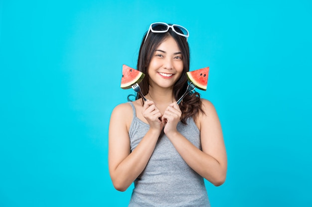 Portrait Attractive Asian young woman holding two Piece slide of watermelon and wearing fashion sunglasses on isolated blue wall, copy space and studio, diet and healthy fruit concept