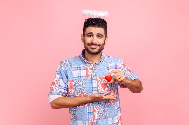 Portrait of attractive bearded man with nimb over head holding small red heart in hands looking at camera with love wearing blue casual shirt Indoor studio shot isolated on pink background