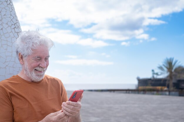 Ritratto di attraente uomo anziano barbuto in piedi all'aperto guardando il telefono cellulare. cielo nuvoloso sullo sfondo, orizzonte sull'acqua