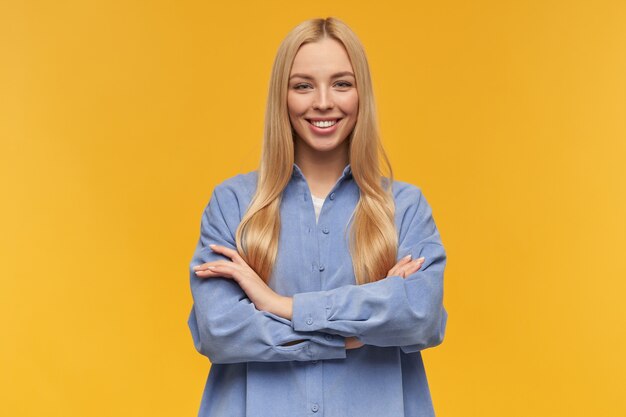 Portrait of attractive, beautiful girl with blond long hair. wearing blue shirt. people and emotion concept. holds arms crossed on a chest. watching at the camera, isolated over orange background