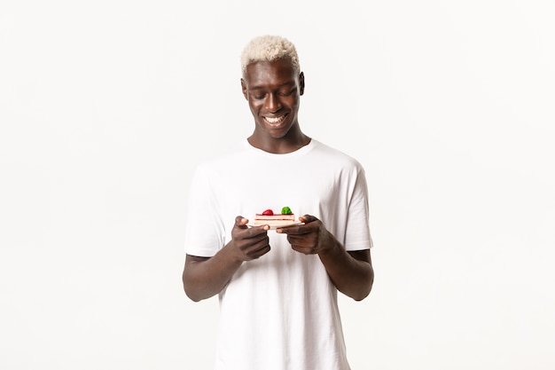 Photo portrait of attractive blond african-american guy, smiling happy and looking at delicious piece of cake