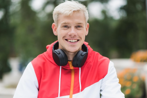 Ritratto di attraente biondo bell'uomo con un sano sorriso a trentadue denti con le cuffie sul collo che si rilassano nel parco della città street fashion style persone stile di vita e concetto di tecnologia