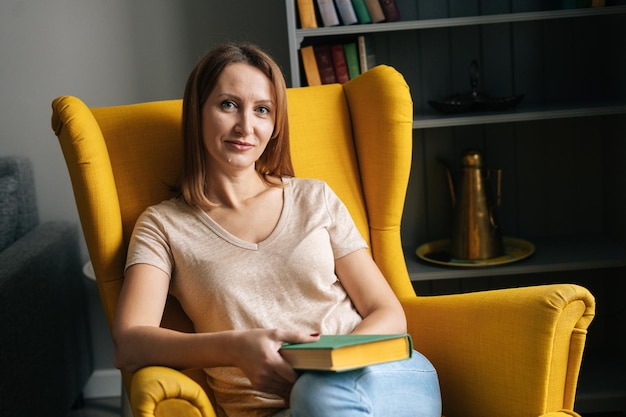 Portrait of attractive blonde woman resting in comfortable yellow chair holding paper book in dark room with modern interior smiling looking at camera